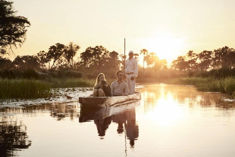 A couple on a romantic safari in Africa seated in a traditional canoe, guided by a standing boatman with a pole, gliding through calm waters surrounded by reeds and trees under a warm, golden sunset