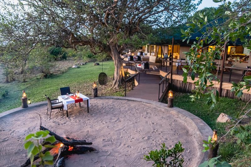 A candlelit dinner beside a small fire pit sits beneath a sprawling tree outside a tented suite, capturing the intimate atmosphere found at Luxury safari lodges in Tanzania.