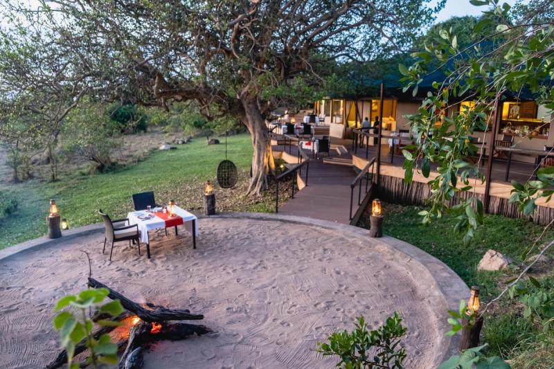 A candlelit dinner beside a small fire pit sits beneath a sprawling tree outside a tented suite, capturing the intimate atmosphere found at Luxury safari lodges in Tanzania.