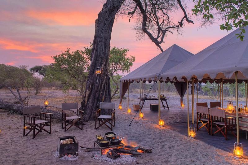 A mobile tented camp glows with lanterns at dusk, featuring canvas dining tents, director's chairs, and a firepit where food is cooked over open coals.