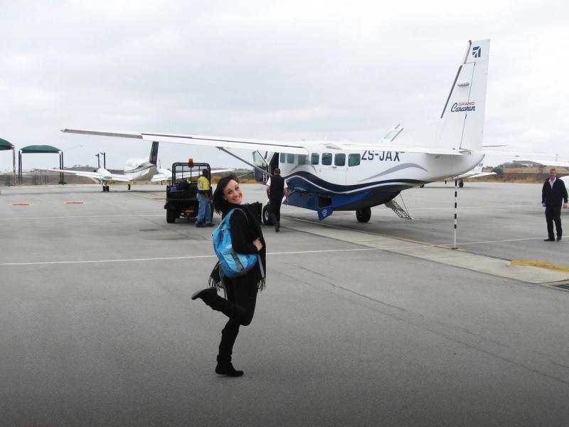 A Rhino Africa travel consultant with a blue backpack smiles on the tarmac beside a small propeller plane as staff prepare for boarding.