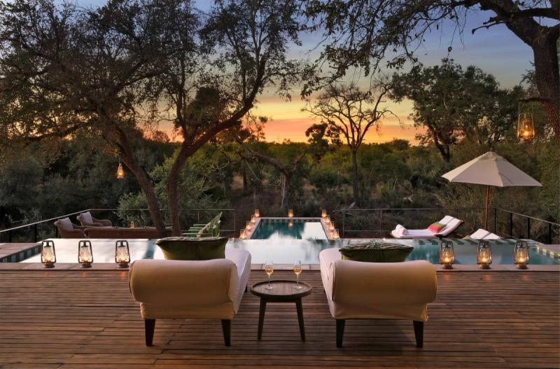 An elegant pool deck surrounded by lantern-lit trees, overlooking a serene wilderness at sunset with inviting loungers and cosy seating