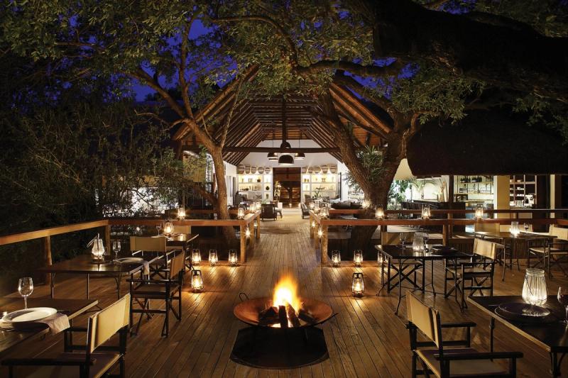 Mauritius and safari holiday: a beautifully lit safari lodge deck, nestled among trees, features elegant dining tables, glowing lanterns, and a central fire pit under the night sky.