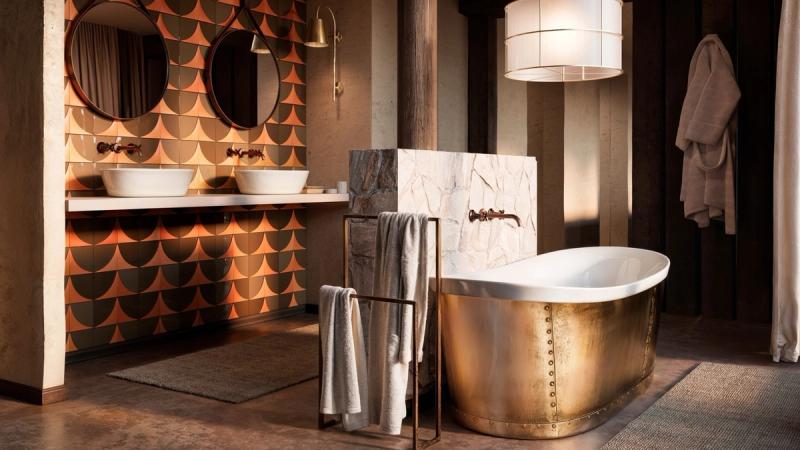 A warmly lit bathroom features patterned tiles, twin basins, and a brass-clad bathtub with soft towels draped nearby.