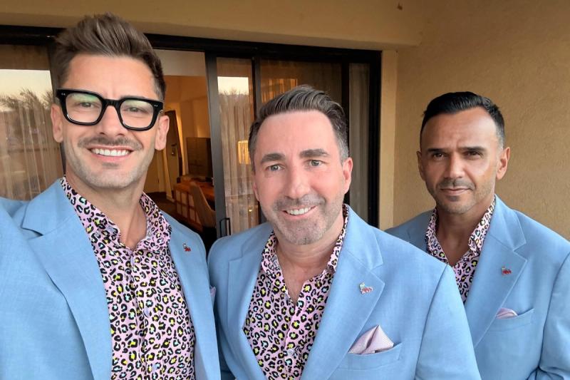 David, Dawid, and Barry wearing matching pastel-blue blazers and pink leopard-print shirts stand smiling together on a balcony.
