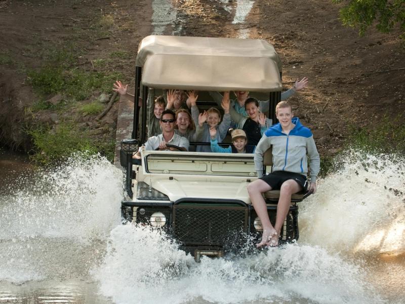 A family-filled safari vehicle splashes through a water crossing as cheerful passengers wave, with one adventurous teen sitting on the vehicle's front edge.
