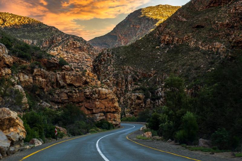 A smooth, winding road cuts through dramatic mountainous terrain, bordered by rugged cliffs and lush greenery.