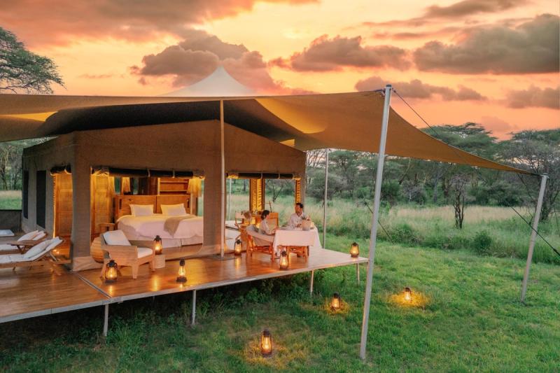 A couple dines on the deck of one of Africa’s most luxurious mobile tented camps, framed by lantern light and a glowing Serengeti sunset.