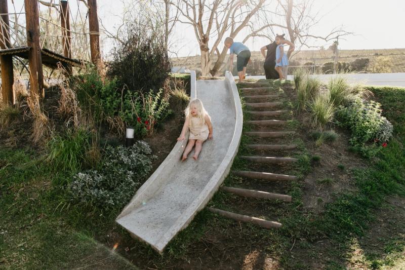 A barefoot toddler slides down a garden play structure while other children wait their turn, capturing how the best destinations for kids can offer safe, joyful spaces to roam.