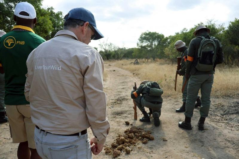 Tracker Academy team studies a dung pile and spoor on a dirt track during field training, while armed rangers stand watch in the bush.