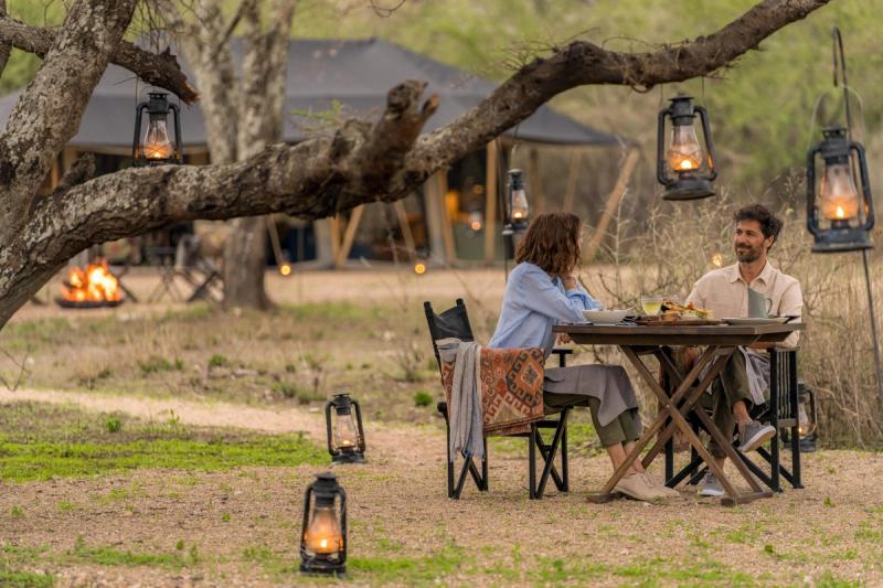 A couple enjoys a lantern-lit meal beneath a tree at one of Africa’s mobile tented camps, with canvas structures and a glowing firepit visible in the background.