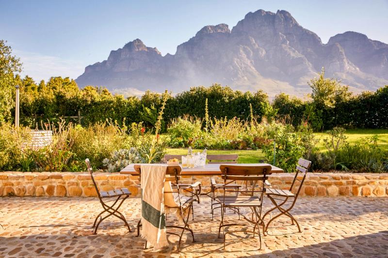 An outdoor wooden dining table set for two sits on a sun-warmed stone terrace, framed by lush gardens and dramatic mountain peaks, capturing the quiet romance of a South African honeymoon.