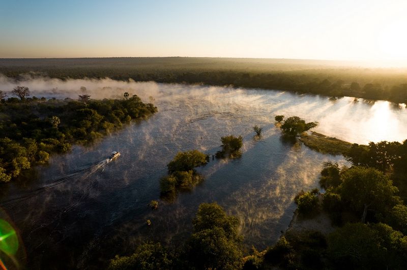 A small boat glides through golden mist on the Zambezi River at sunrise, upstream from the best side of Victoria Falls.