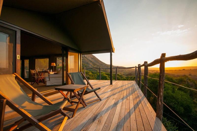 A sun-flooded wooden safari lodge deck with a table and loungers looking out over the wilderness during sunrise