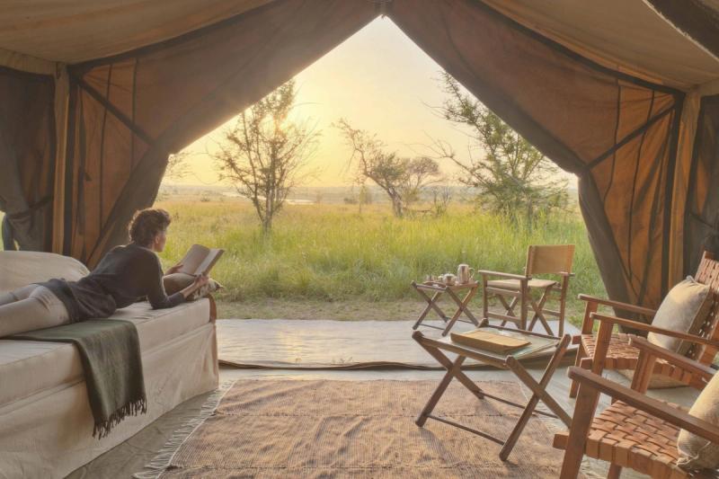 A woman lounges with a book in one of Africa’s mobile tented camps, facing open canvas flaps and a golden view of the wilderness beyond.