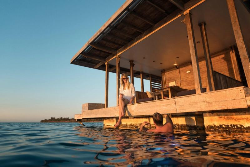 A couple relaxes at a luxurious overwater retreat, with one enjoying the warm ocean waters while the other savours the sunset on the deck
