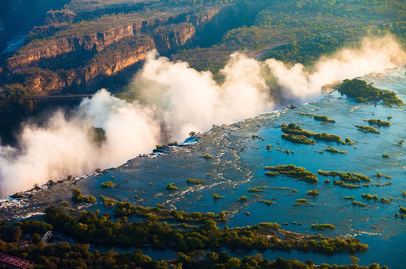 Aerial view of water surging toward the cliff edge, with thick mist rising above the best side of Victoria Falls at peak flow.