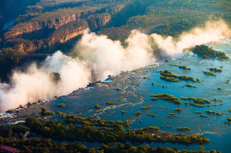 Aerial view of water surging toward the cliff edge, with thick mist rising above the best side of Victoria Falls at peak flow.