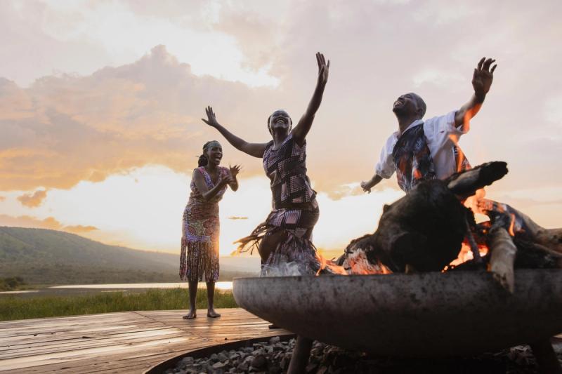 Three barefoot dancers perform around a glowing firepit at sunset, their hands raised in joy against a lake and mountain backdrop.