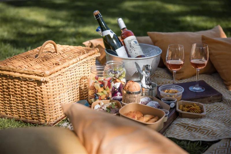 picnic basket, wine, delectable treats, and cushions under oak trees on a lush lawn