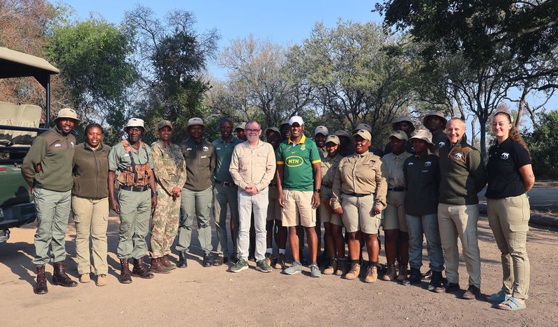 Group photo of Tracker Academy trainers and trainees standing outdoors in field uniforms at Kruger National Park, smiling beside a safari vehicle.