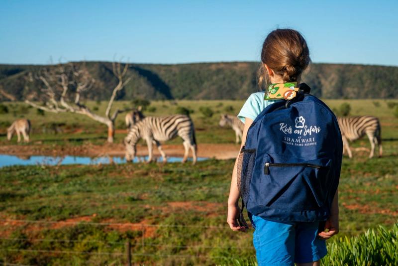 A young girl wearing a “Kids on Safari” backpack gazes at a small herd of zebras drinking at a waterhole, set against a backdrop of green plains and rolling hills.