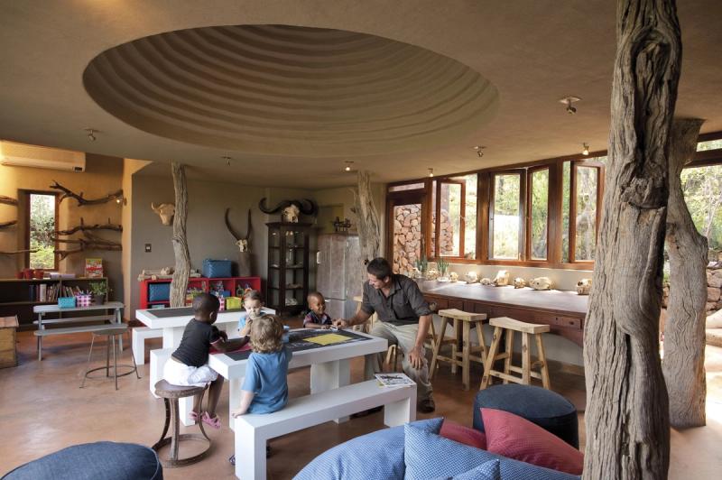 An interactive kids' playroom with tables, stools, and a variety of educational items, where children engage in activities alongside a guide, surrounded by decor inspired by the African bush.