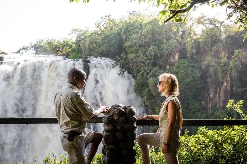 A guide and traveller stand face-to-face beside a railing, with roaring water from the best side of Victoria Falls cascading behind them.