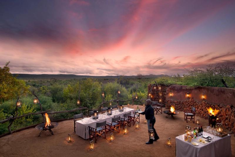 A beautifully set outdoor dining area, illuminated by lanterns and a central fire pit, overlooks a serene bush landscape at sunset, with a staff member preparing the tables.