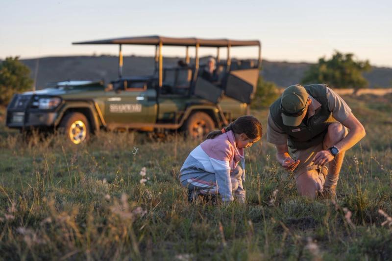 A young girl and a safari guide crouch together in the grassy plains of a malaria-free game reserve, inspecting a small find on the ground with focused curiosity.