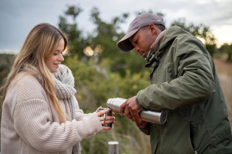 A safari guide in a green jacket pours a warm drink from a thermos for Katharina wrapped in a cosy sweater during her babymoon safari.