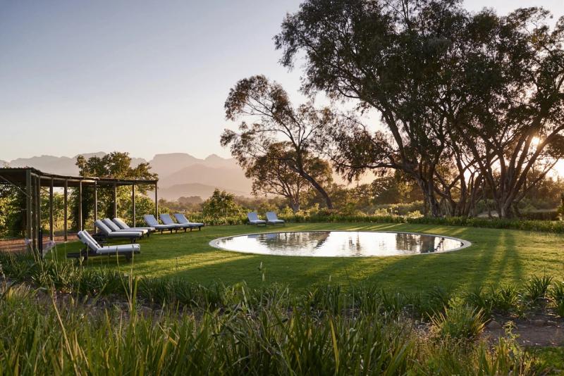 A tranquil circular swimming pool set on manicured lawn, framed by ancient trees and distant mountains, captures the unhurried elegance of a South African honeymoon.