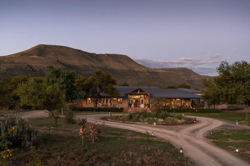 A scenic view of Samara Karoo Lodge nestled at the foot of a mountain, surrounded by natural vegetation at dusk.