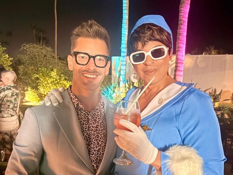 Dawid in a light blue blazer poses with a smiling drag performer dressed in a retro blue outfit and white sunglasses, holding a cocktail.