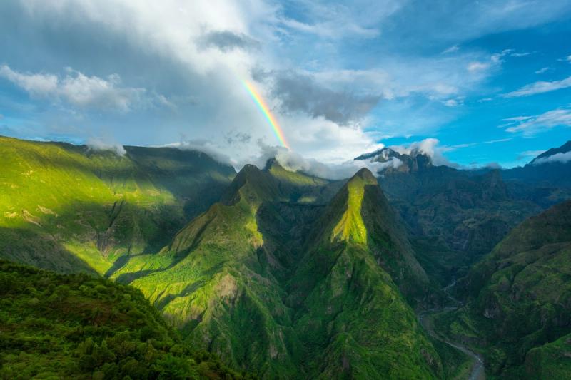 Lush green mountains rise dramatically beneath a bright sky with a vivid rainbow stretching through scattered clouds
