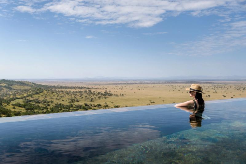 A woman in a straw hat relaxes in an infinity pool overlooking the vast Serengeti plains, with scattered acacia trees and distant hills under a clear blue sky.