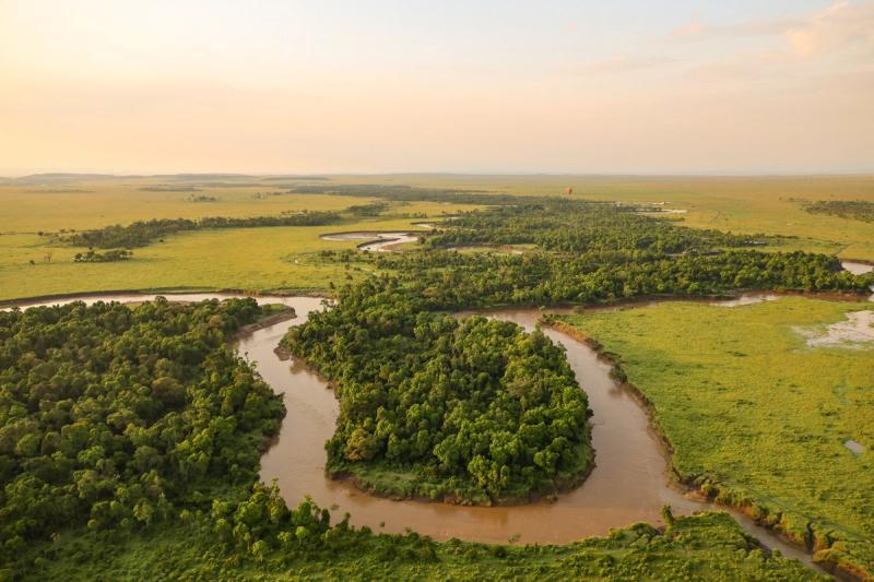 A winding river snakes through lush green plains and dense forest under soft golden light, with a hot air balloon faintly visible in the distance