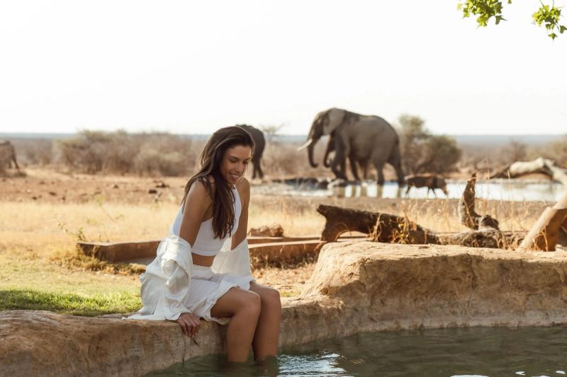 A woman sits by a pool dipping her feet in the water, with elephants drinking at a nearby watering hole in a dry, open landscape.