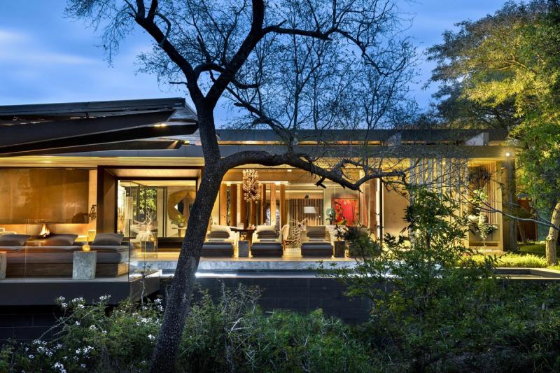 A softly lit, modern safari lodge glowing at dusk among trees beside a nearby water source, capturing the quiet elegance of lodges with the best waterholes.