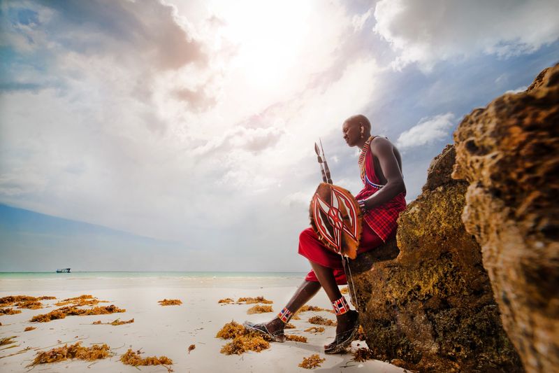 A Maasai warrior sits facing the ocean, framed by rugged rocks and calm white sands