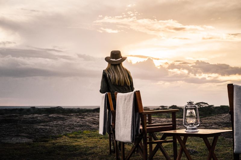 A woman in a safari hat sits facing the horizon at sunset, with empty wooden chairs and a lantern nearby on the grass