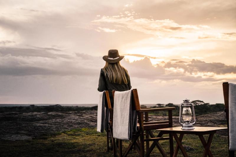 A woman in a safari hat sits facing the horizon at sunset, with empty wooden chairs and a lantern nearby on the grass