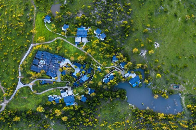 An aerial view of a modern safari lodge spread across green bushveld, with villas clustered around a prominent waterhole, revealing how lodges with the best waterholes are thoughtfully shaped by water and space.