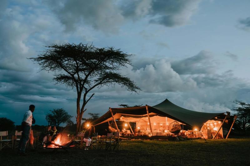 A glowing canvas pavilion, lit from within, anchors this mobile tented camp in the Serengeti as guests gather around a fire under the evening sky.