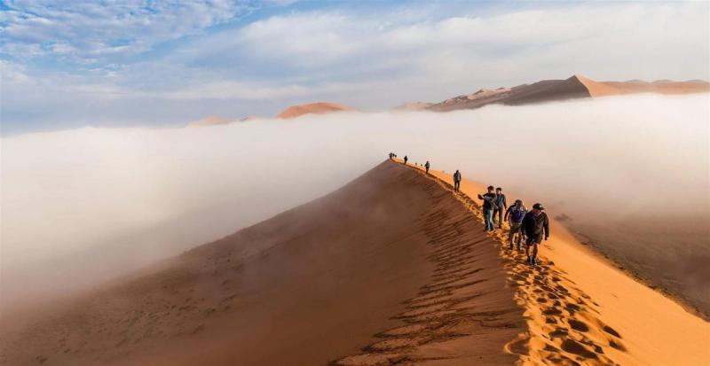 A group of travellers walk along a narrow ridge of a towering red sand dune surrounded by mist and distant desert peaks