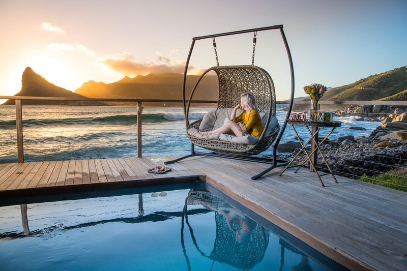 A woman sits on a hanging chair overlooking the ocean and mountains at one of the luxury hotels in Cape Town