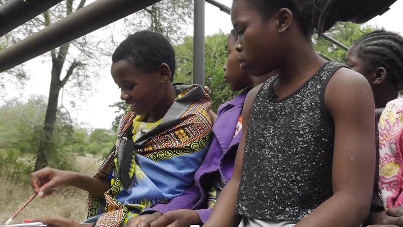Girls sit together on a vehicle bench, one drawing in her notebook during a Gorongosa safari.