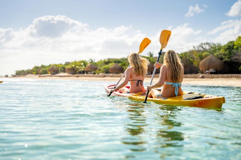 Two young adults kayak through crystal-clear water toward a tropical shore, proving that luxury island destinations for kids-turned-grownups still bring the adventure.