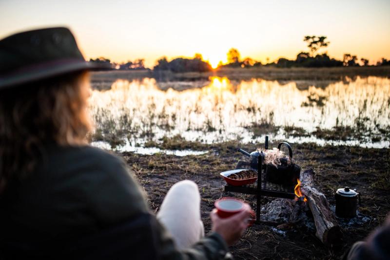 A guest at one of Africa’s mobile tented camps sits beside a fire with coffee and rusks at sunrise, overlooking a still floodplain.