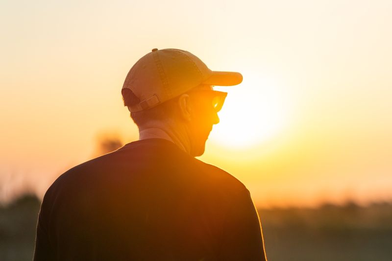 David wearing a cap and sunglasses faces a glowing sunset, his profile softly lit by the warm orange light
